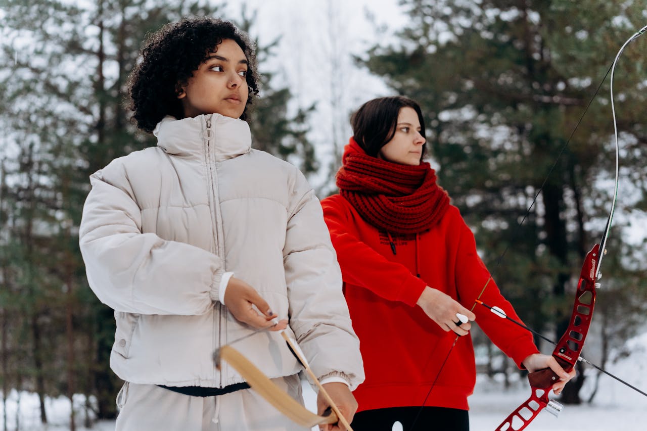 about-02 Two women practice archery in a snowy forest, focusing on their technique amidst the winter scenery.